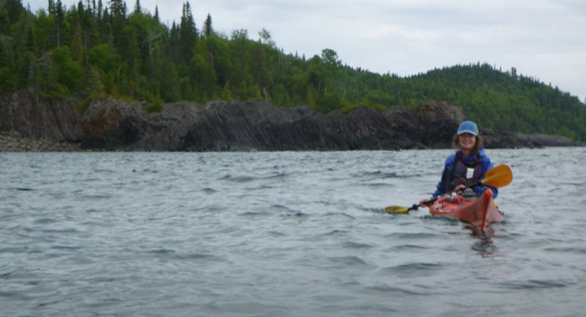 a woman wearing a life jacket paddles a red canoe toward the camera. behind her is a tree-lined shore.
